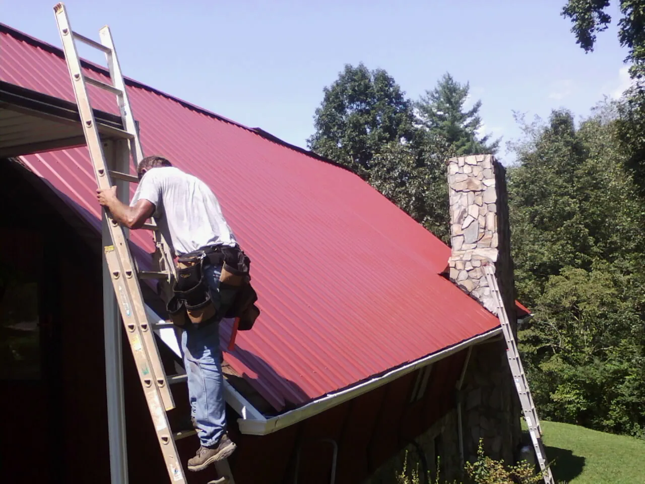 Roofer on a ladder beside a red metal roof and stone chimney — Soffit & Fascia Repair by Roofing Brothers in Kannapolis